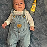 baby, infant, denim_overalls, teddy_bear_patch, blanket, dinosaur_pattern, bottle, chamomile_spray, laying_down, curious_expression, clothing, footwear_absent, hand, face, head, skin, indoors, floor, soft_texture, cute