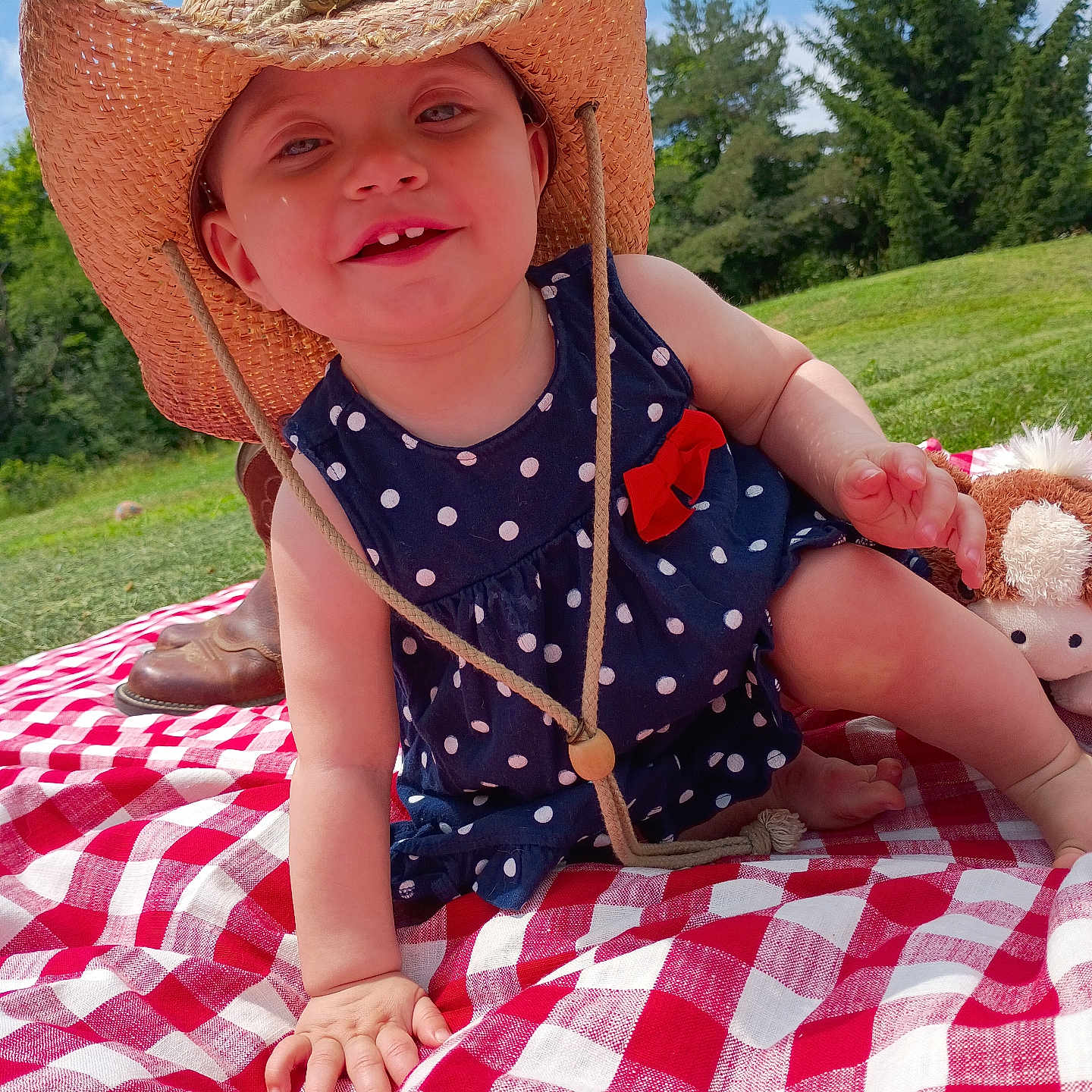Jocelyn is registered to the contest to win money with this photo: baby, child, cowboy_hat, polka_dot, dress, red_bow, picnic_blanket, outdoor, grass, trees, smiling, happy, sunny, toy, stuffed_animal, hand, face, blue_sky, foot, nature