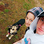 Harley is registered to the contest to win money with this photo: person, dog, hoodie, outdoor, grass, casual_clothing, pet, leash, canine, looking_up, portrait, two_people, young_adult, casual, brown_dog, black_and_tan_dog, curious_dog, headwear, cap, nature