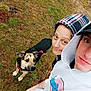 person, dog, hoodie, outdoor, grass, casual_clothing, pet, leash, canine, looking_up, portrait, two_people, young_adult, casual, brown_dog, black_and_tan_dog, curious_dog, headwear, cap, nature