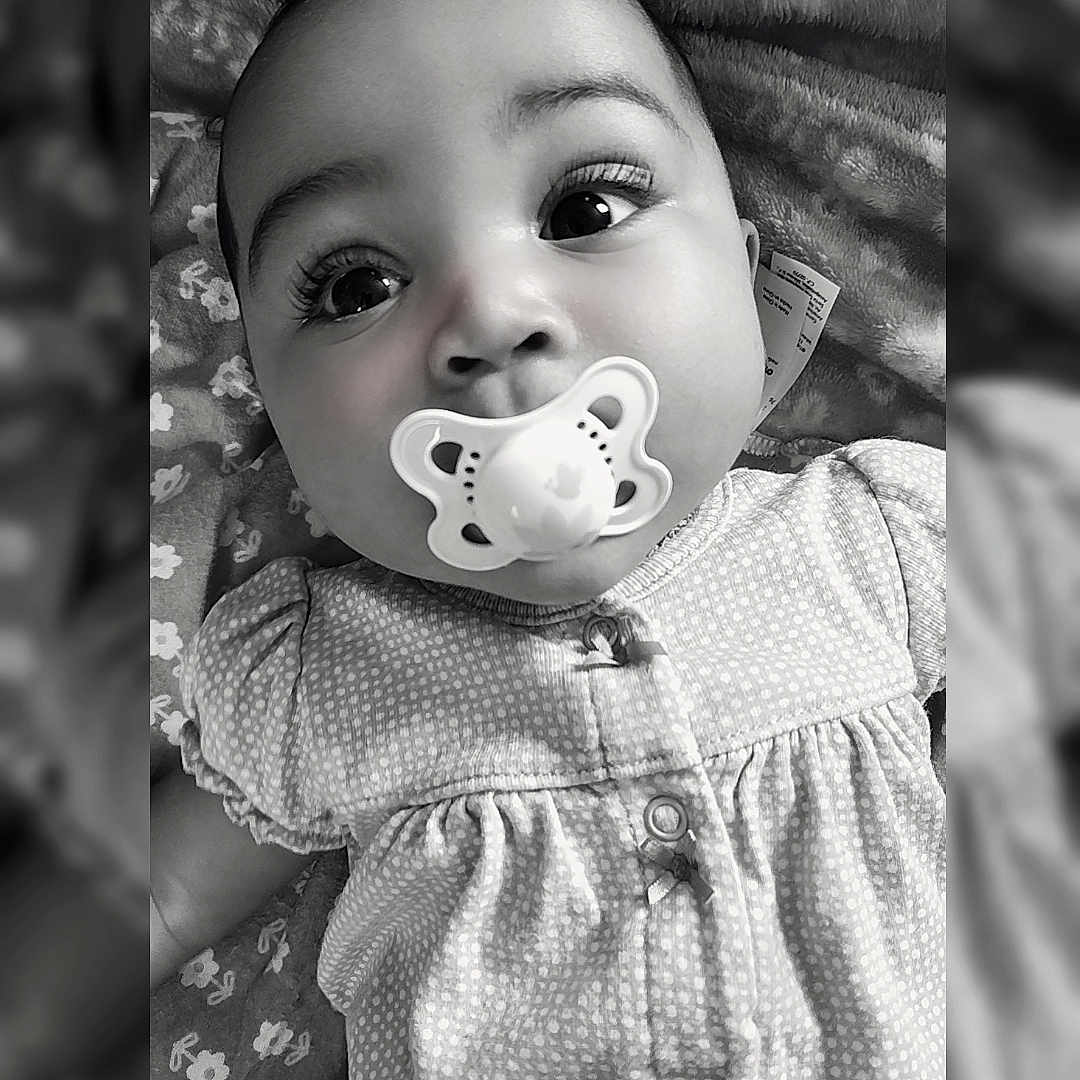 Rae is registered to the contest to win money with this photo: adorable, baby, baby_girl, black_and_white, blanket, child, close_up, clothing, cute, dress, expression, eyes, face, infant, lying_down, pacifier, pattern, portrait, soft_texture, young