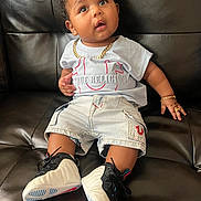 Suriel is registered to the contest to win money with this photo: baby, bracelet, child, curious, cute, expression, fashion, footwear, furniture, indoors, jewelry, leather_couch, necklace, person, portrait, shorts, sitting, sneakers, tshirt, young