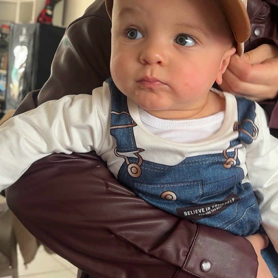Ayden a rejoint le concours — aidez-le/la à gagner de superbes lots ! baby, child, cap, denim_overalls, adult, indoor, person, holding, curious, expression, maroon_jacket, face, headwear, clothing, home, sitting, looking_away, casual, cute, portrait