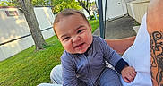 Johdi is registered to the contest to win money with this photo: baby, child, striped_onesie, outdoor, grass, tree, person, tattoo, adult, lawn, mobile_home, sunny, sidewalk, blue_sky, smiling, cute, face, hand, footed_pajamas, casual