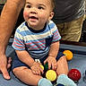 toddler, child, pool_table, billiard_balls, blue_socks, striped_shirt, shorts, indoor, smiling, person, adult_arm, playing, colorful, seated, fun, curious, happy, game_room, family, support