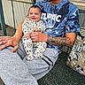 baby, man, smile, tshirt, tattoo, bench, cushion, pants, onesie, animal_print, outdoor, metal_bench, person, holding, affection, happy, casual_clothing, sitting, portrait, parent_child