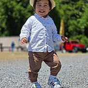 Milán David is registered to the contest to win money with this photo: toddler, child, hat, white_shirt, brown_pants, sneakers, outdoor, smiling, walking, gravel, daylight, greenery, trees, red_vehicle, casual_clothing, happy, young_child, sunny, portrait, nature