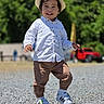 toddler, child, hat, white_shirt, brown_pants, sneakers, outdoor, smiling, walking, gravel, daylight, greenery, trees, red_vehicle, casual_clothing, happy, young_child, sunny, portrait, nature