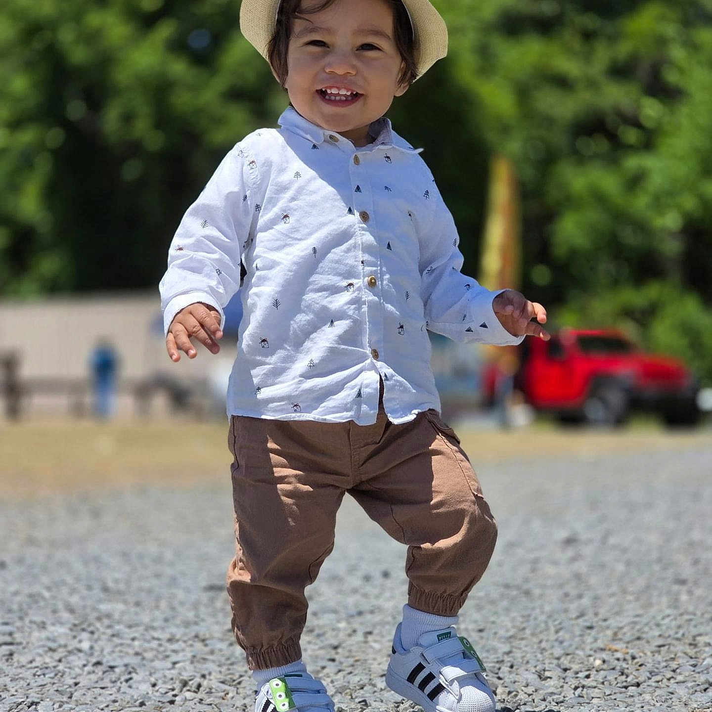 Milán David is registered to the contest to win money with this photo: toddler, child, hat, white_shirt, brown_pants, sneakers, outdoor, smiling, walking, gravel, daylight, greenery, trees, red_vehicle, casual_clothing, happy, young_child, sunny, portrait, nature