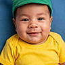 baby, child, portrait, smiling, happy, green_hat, mouse_ears, yellow_shirt, blue_background, close_up, face, eyes, indoors, studio_portrait, infant, sitting, colorful, clothing, centered_composition, adorable