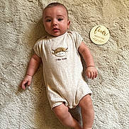 Evan a rejoint le concours — aidez-le/la à gagner de superbes lots ! baby, infant, child, onesie, plush_rug, milestone_marker, four_months, wooden_plaque, cute, smiling, lying_down, soft_texture, beige, portrait, indoor, baby_clothing, face, hand, foot, milestone