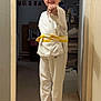 child, karate_gi, yellow_belt, martial_arts, barefoot, smiling, indoor, hallway, floor, doorway, pose, young, person, clothing, happy, pants, short_hair, white_clothing, playful, karate