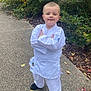 child, martial_arts_uniform, karate_gi, smiling, standing, outdoor, nature, greenery, bushes, pathway, autumn_leaves, black_shoes, pose, young_child, casual, confident, daylight, person, cute, happy