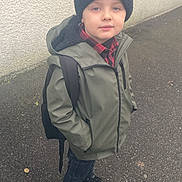 Kylan participe au concours pour gagner de l'argent avec cette photo : backpack, boy, casual, child, clothing, cold_weather, face, footwear, green_jacket, headwear, looking_at_camera, outdoor, pavement, portrait, sidewalk, standing, street, wall, winter_hat, young