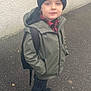 child, boy, winter_hat, green_jacket, backpack, sidewalk, street, wall, pavement, standing, outdoor, casual, clothing, footwear, portrait, face, looking_at_camera, young, cold_weather, headwear