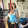 blue_skirt, certificates, chair, child, computer_chair, curtain, figurines, floor, folded_clothes, girl, home, peace_sign, pink_socks, room, shelf, standing, table, toys, white_top, window