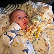 Denton is registered to the contest to win money with this photo: baby, infant, onesie, shark_print, crocheted_toy, octopus_toy, blanket, cozy, cute, child, lying_down, soft_texture, indoor, curious, blue, yellow, white, patterned_clothing, newborn, comfort