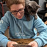 person, cat, glasses, hoodie, card_game, table, indoor, wooden_table, gray_cat, playing_cards, sitting, pet, shoulder, focused, casual_clothing, home, furniture, relaxed, game, curious