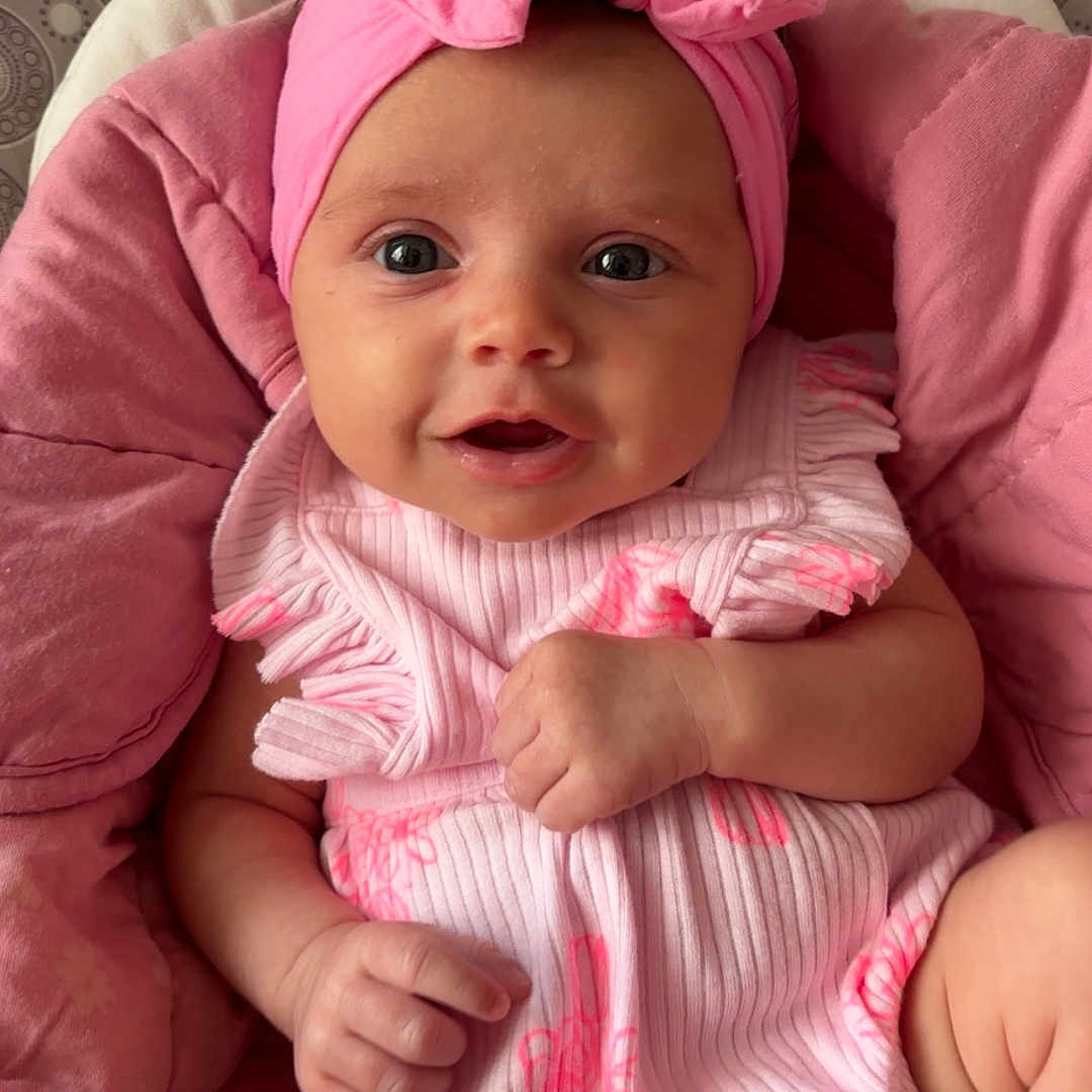 Celine is registered to the contest to win money with this photo: baby, bow, child, closeup, clothing, comfortable, cushion, cute, face, hand, headband, indoors, infant, legs, patterned_background, pink, portrait, skin, smiling, soft_texture