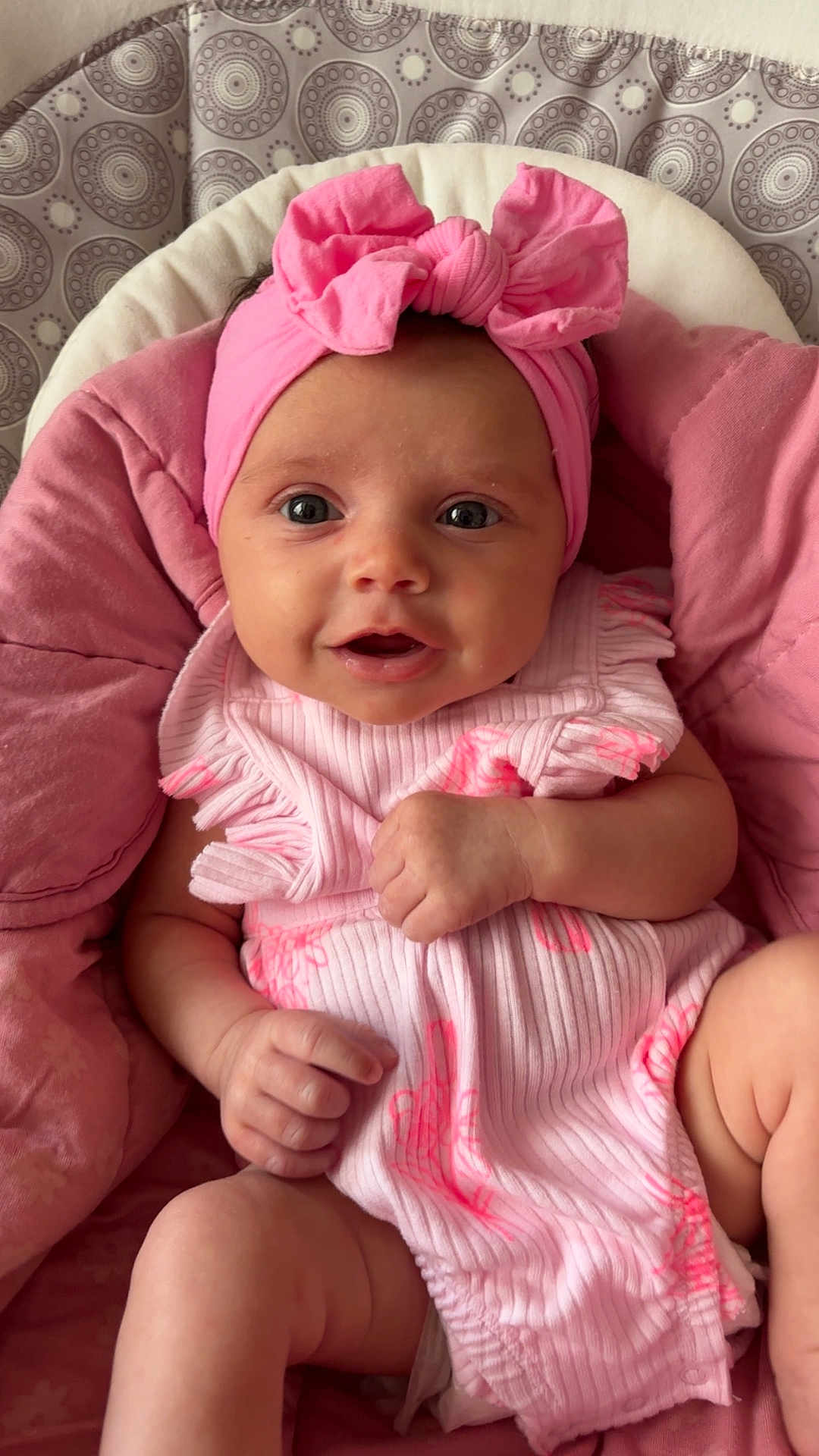 Celine is registered to the contest to win money with this photo: baby, infant, pink, headband, bow, clothing, cushion, patterned_background, smiling, face, skin, hand, legs, cute, portrait, child, indoors, soft_texture, closeup, comfortable