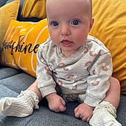 Maisie is registered to the contest to win money with this photo: baby, infant, sitting, couch, pillow, yellow_pillow, gray_couch, sweater, rainbow_pattern, white_socks, frills, indoor, child, cute, face, eyes, mouth, hands, legs, soft_lighting