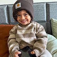 Mayronn a rejoint le concours — aidez-le/la à gagner de superbes lots ! child, smiling, beanie, brown, curly_hair, sweater, striped, smartphone, couch, cushion, indoor, happy, person, seated, cozy, casual_clothing, furniture, portrait, young_child, playful