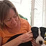 air_conditioner, black_and_white, casual, curious, curtains, dog, furniture, gentle, hand, holding, home, indoor, light, orange_shirt, pet, portrait, puppy, smiling, window, woman