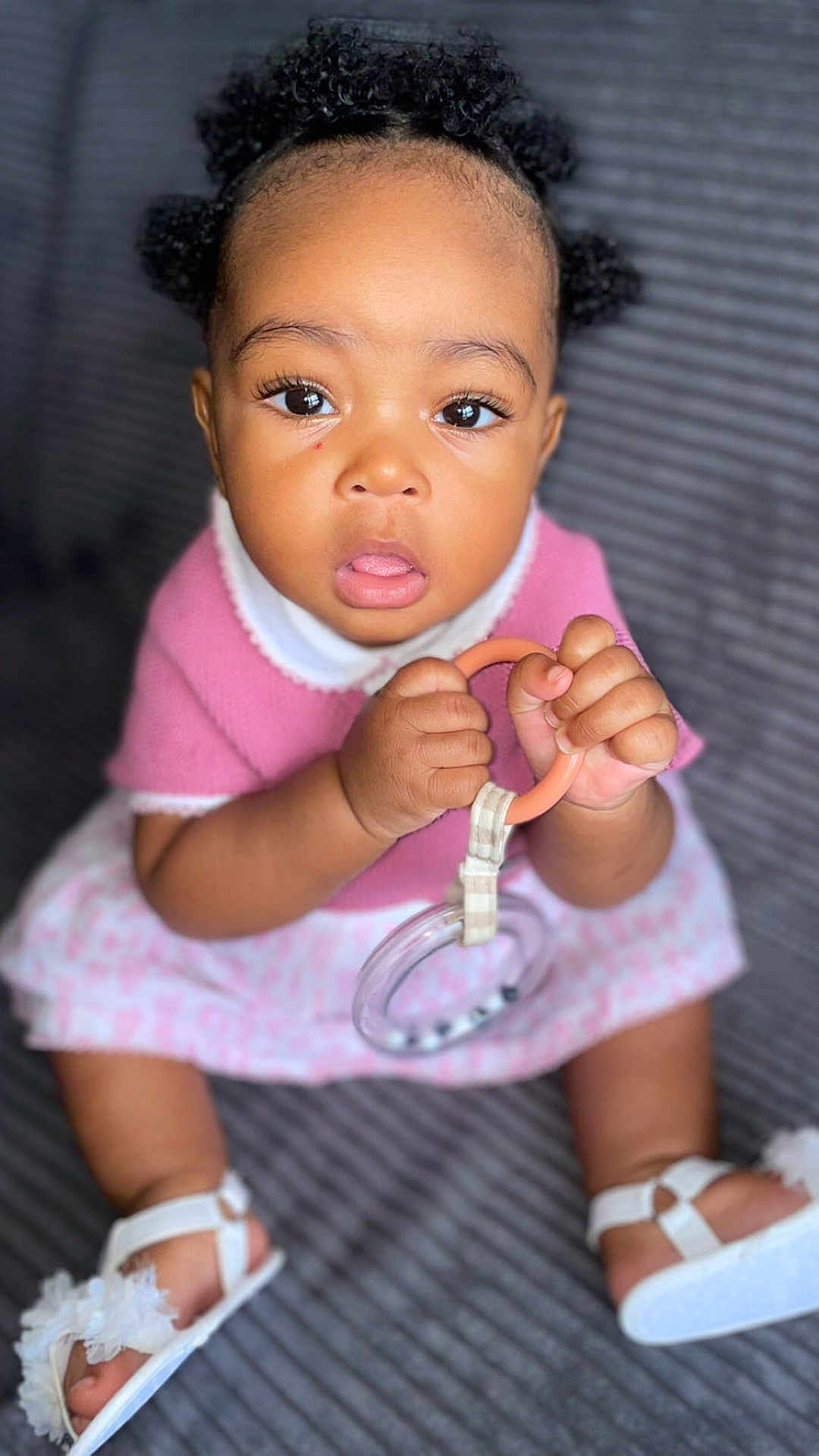 Nellya a rejoint le concours — aidez-le/la à gagner de superbes lots ! baby, child, curly_hair, pink_clothing, toy, ring, white_sandals, open_mouth, wide_eyes, sitting, indoor, close_up, cute, infant, hands, face, footwear, person, portrait, texture