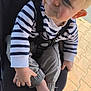 toddler, child, smile, striped_shirt, beige_pants, barefoot, stroller, seatbelt, outdoor, pavement, footwear, hand, face, blue_eyes, blond_hair, clothing, person, sitting, cute, sunlight