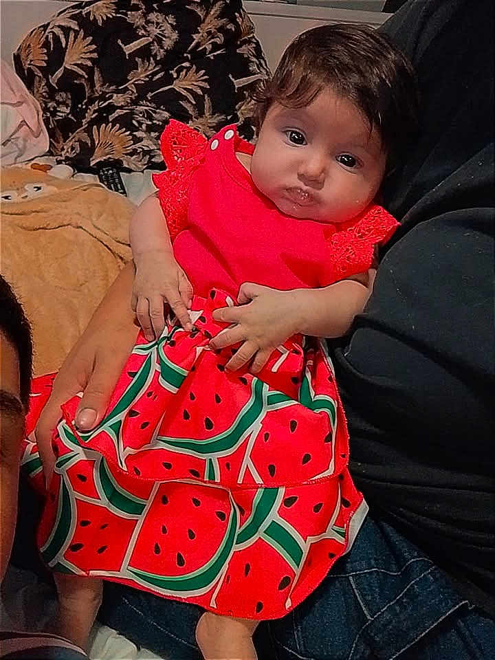 Kataleya participe au concours pour gagner de l'argent avec cette photo : baby, infant, child, watermelon_dress, red_clothing, hands, adult_arm, couch, blanket, patterned_fabric, dark_hair, big_eyes, portrait, indoors, holding, cute, sitting, family_moment, soft_lighting, plush_toy