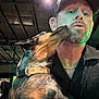 affection, animal, beard, blur, casual_clothing, ceiling, chain_necklace, closeup, collar, dog, friendship, hat, human, indoor, interaction, licking, light, man, pet, portrait