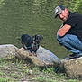 animal, calm, cap, casual_clothing, crouching, dachshund, daytime, dog, grass, greenery, leash, man, nature, outdoor, pet, river, rock, scenic, small_dog, water