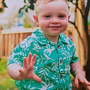 Freddie is registered to the contest to win money with this photo: toddler, child, smiling, hand, green_shirt, floral_pattern, outdoor, garden, fence, happy, portrait, person, cute, young_child, short_hair, summer, nature, tree, casual_clothing, daylight