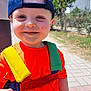 Freddie is registered to the contest to win money with this photo: toddler, child, smile, cap, orange_shirt, straps, outdoor, sunny, daylight, happy, person, sidewalk, tree, fence, bracelet, face, casual_clothing, young_child, portrait, background_blur