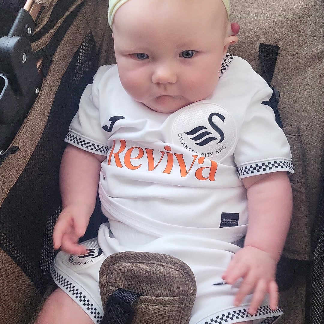 Ivy May is registered to the contest to win money with this photo: baby, bow, child, chubby, closeup, cute, expression, fabric, headband, indoor, infant, person, seated, soccer, sportswear, straps, stroller, swansea_city, white_clothing, yellow_accessory