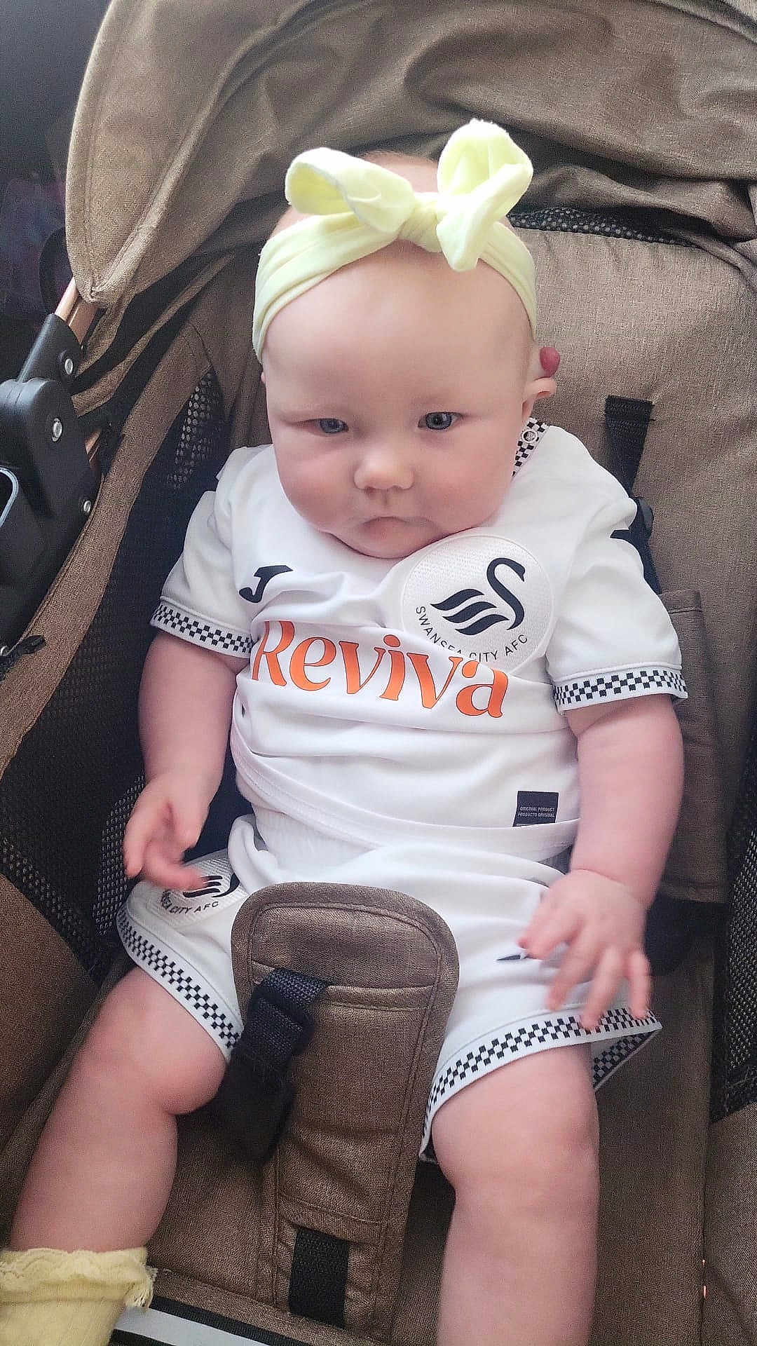 Ivy May is registered to the contest to win money with this photo: baby, child, infant, stroller, headband, bow, sportswear, soccer, swansea_city, white_clothing, yellow_accessory, cute, chubby, expression, seated, indoor, fabric, straps, person, closeup