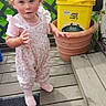 baby, candid, casual, child, curious, daylight, double_buns, foot, greenery, outdoor, painted_nails, person, pink_outfit, plant_pot, standing, tattoo, tidy_cats, toddler, wooden_porch, yellow_container