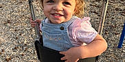 Wrenlee is registered to the contest to win money with this photo: toddler, child, swing, playground, smile, denim_overalls, pink_shirt, chains, wood_chips, outdoor, daylight, happy, curly_hair, holding, park, fun, person, young_child, clothing, nature
