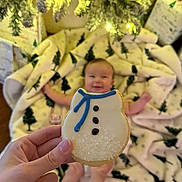 Josephine joined the competition — help win amazing prizes! baby, cookie, hand, blanket, christmas_tree_pattern, holiday_lights, gift_wrap, smile, blurred_background, snowman_cookie, infant, festive, indoor, celebration, cozy, decorations, warm_lighting, person, food, sweet