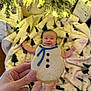 baby, cookie, hand, blanket, christmas_tree_pattern, holiday_lights, gift_wrap, smile, blurred_background, snowman_cookie, infant, festive, indoor, celebration, cozy, decorations, warm_lighting, person, food, sweet