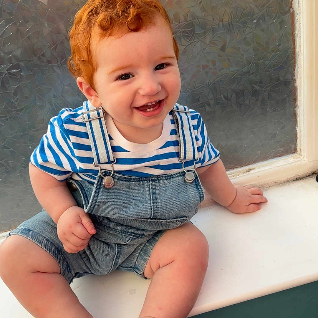 Liam is registered to the contest to win money with this photo: baby, casual_clothing, child, curly_hair, cute, denim_overalls, foot, glass, hand, happy, indoor, person, portrait, red_hair, skin, smiling, striped_shirt, toddler, window, windowsill