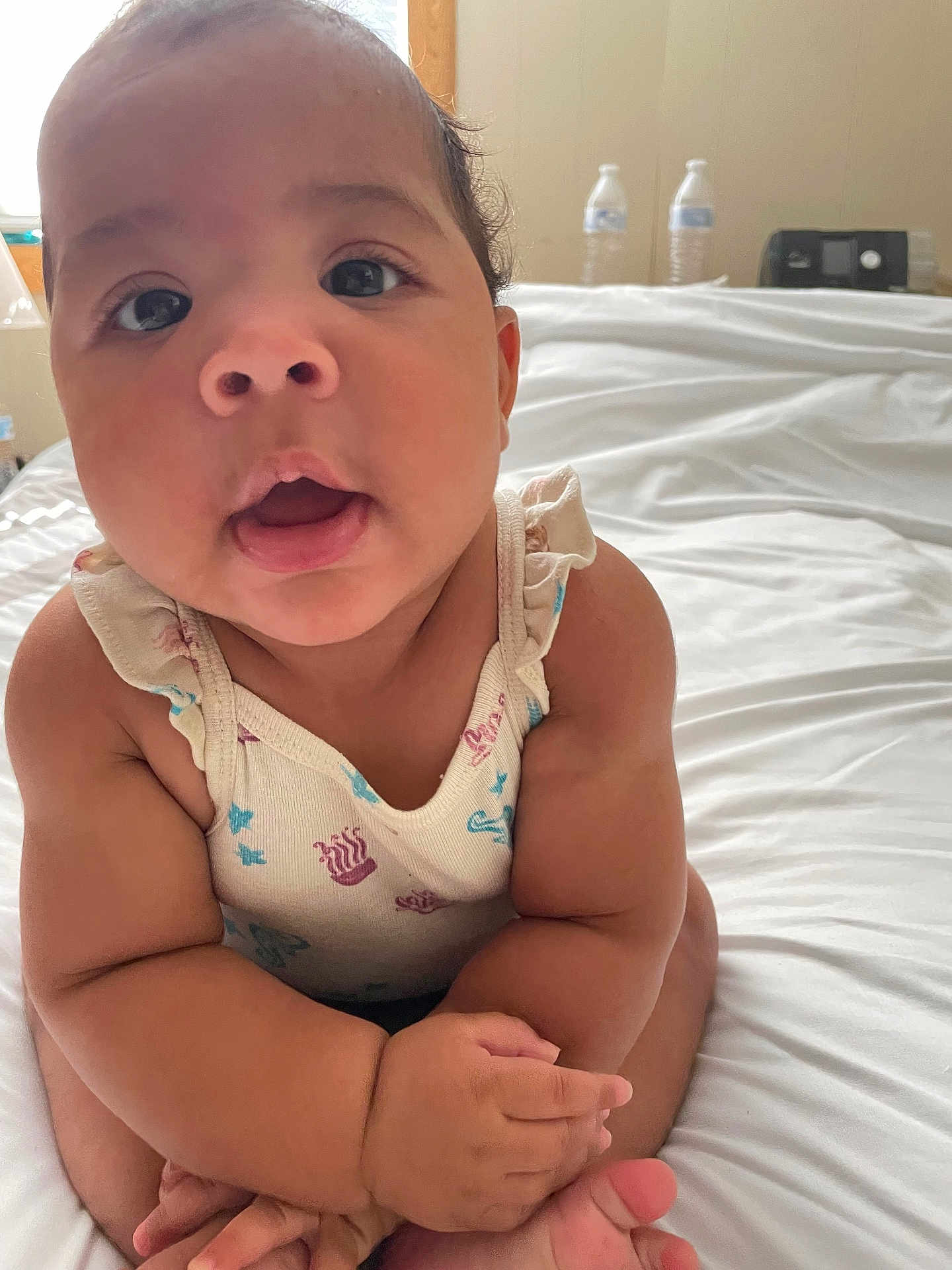 Maylee joined the competition — help win amazing prizes! baby, infant, child, cute, face, eyes, mouth, arms, hands, legs, sitting, bed, white_bedspread, clothing, patterned_outfit, indoors, room, water_bottle, curious, portrait