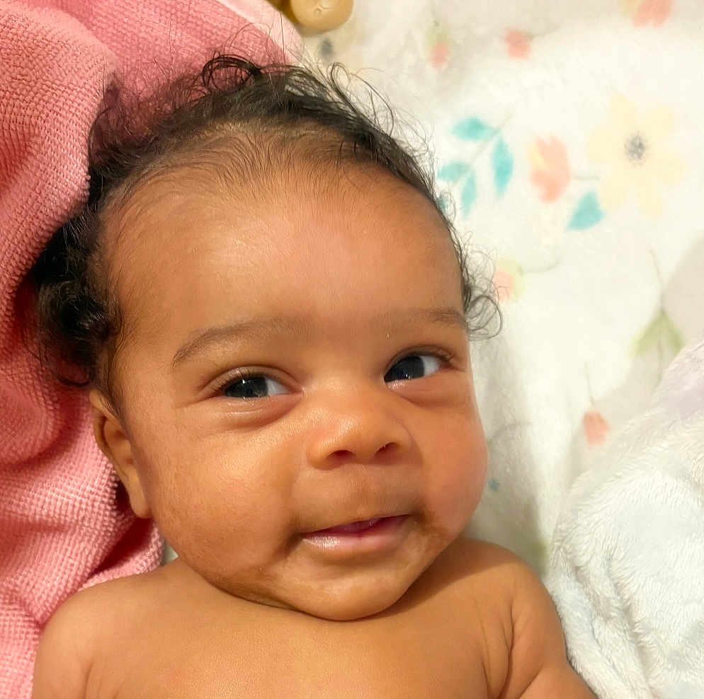 Kasani is registered to the contest to win money with this photo: baby, infant, face, smile, eyes, skin, hair, blanket, pink, fabric, soft, cute, closeup, portrait, child, newborn, lying_down, indoors, expression, adorable