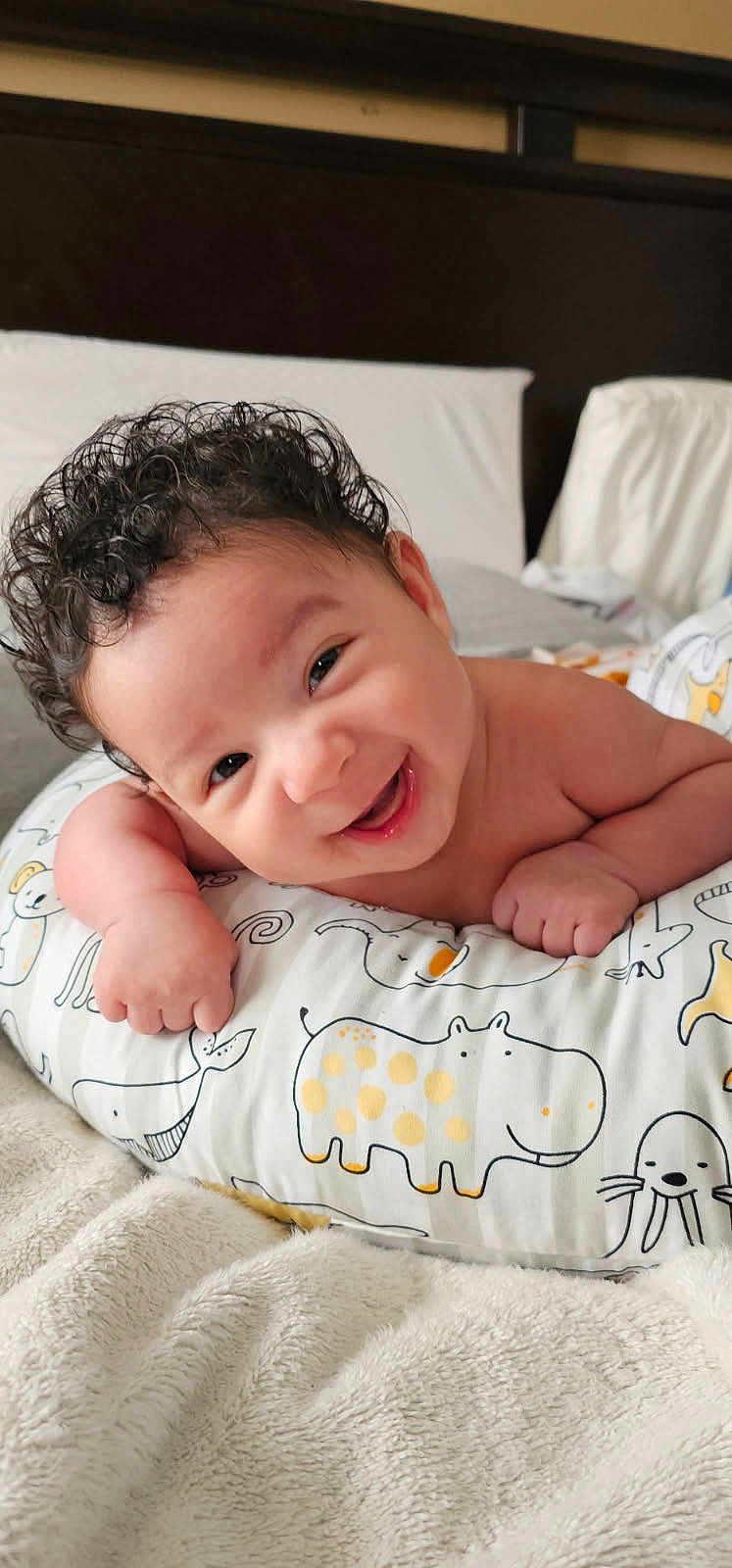 Zayden joined the competition — help win amazing prizes! baby, smiling, curly_hair, pillow, animal_pattern, bedroom, blanket, infant, happy, cute, child, resting, indoors, soft_texture, face, person, young, comfort, toddler, expression