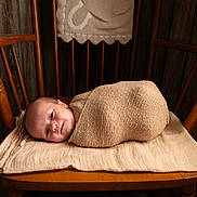 Beckham is registered to the contest to win money with this photo: baby, newborn, wrapped, blanket, wooden_chair, cushion, face, infant, indoors, wood_paneling, cozy, portrait, child, cute, resting, soft_lighting, warm_colors, homedecor, peaceful, smiling
