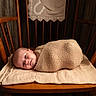 baby, newborn, wrapped, blanket, wooden_chair, cushion, face, infant, indoors, wood_paneling, cozy, portrait, child, cute, resting, soft_lighting, warm_colors, homedecor, peaceful, smiling