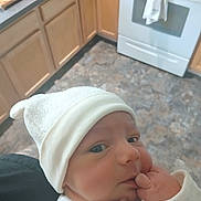 Luke joined the competition — help win amazing prizes! baby, infant, white_hat, kitchen, wooden_cabinets, stove, floor, hand, face, clothing, person, indoor, newborn, curious, soft_light, home, portrait, cute, sleeves, skin