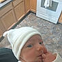 baby, infant, white_hat, kitchen, wooden_cabinets, stove, floor, hand, face, clothing, person, indoor, newborn, curious, soft_light, home, portrait, cute, sleeves, skin