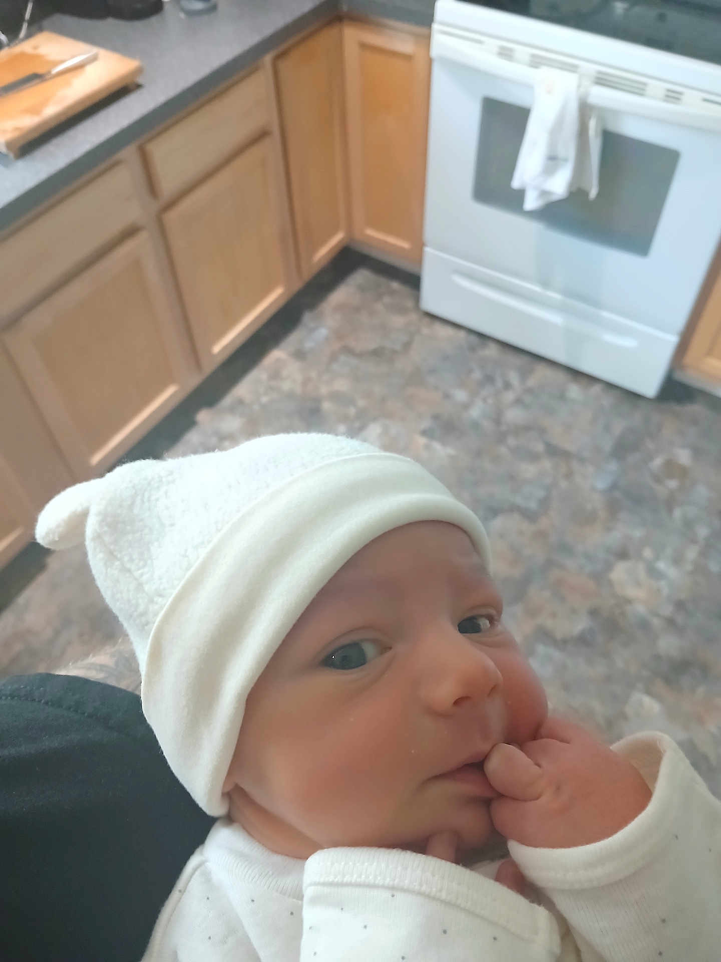 Luke joined the competition — help win amazing prizes! baby, infant, white_hat, kitchen, wooden_cabinets, stove, floor, hand, face, clothing, person, indoor, newborn, curious, soft_light, home, portrait, cute, sleeves, skin