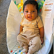 Demarion is registered to the contest to win money with this photo: baby, infant, child, curly_hair, beige_clothing, socks, baby_seat, carpet, playful, tongue_out, cute, indoors, colorful_background, giraffe_pattern, soft_toys, comfort, smiling, portrait, young_child, happy
